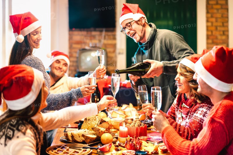 Millenial friends on santa hats celebrating Christmas with champagne and sweets food at log cabin - Winter holidays concept with young people
