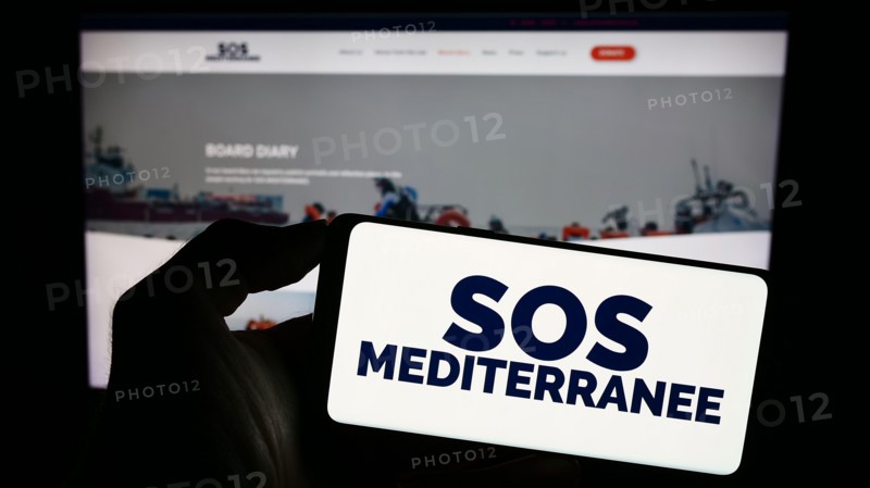 Person holding cellphone with logo of French sea rescue organisation SOS Mediterranee in front of webpage. Focus on phone display.