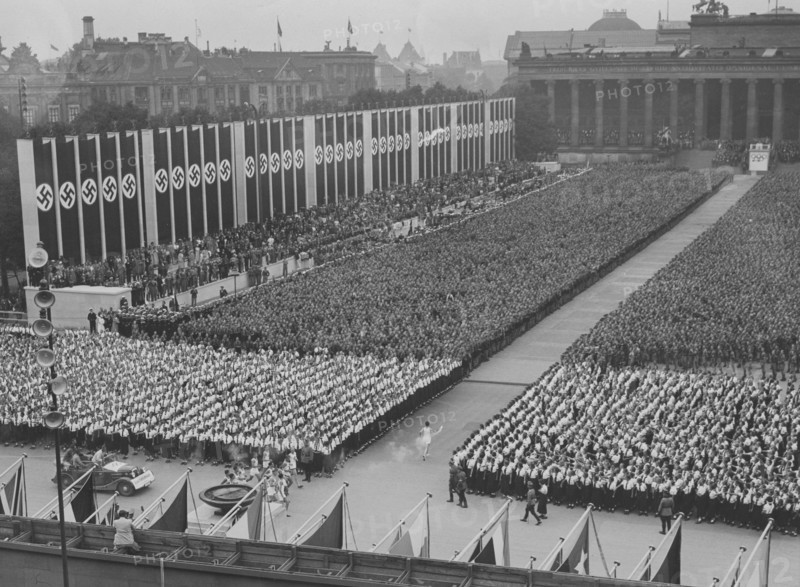 Berlin Olympics 1936. The 1936 Summer Olympics (German: Olympische Sommerspiele 1936), officially known as the Games of the XI Olympiad (German: Spiele der XI. Olympiade) and commonly known as Berlin 1936, were an international multi-sport event held from 1 to 16 August 1936 in Berlin, Germany. Pictured the olympic flame being transported to the Olympiastadion in Berlin by runners, this ceremony is called Olympic torch relay. The nazi flags are seen hanging there.