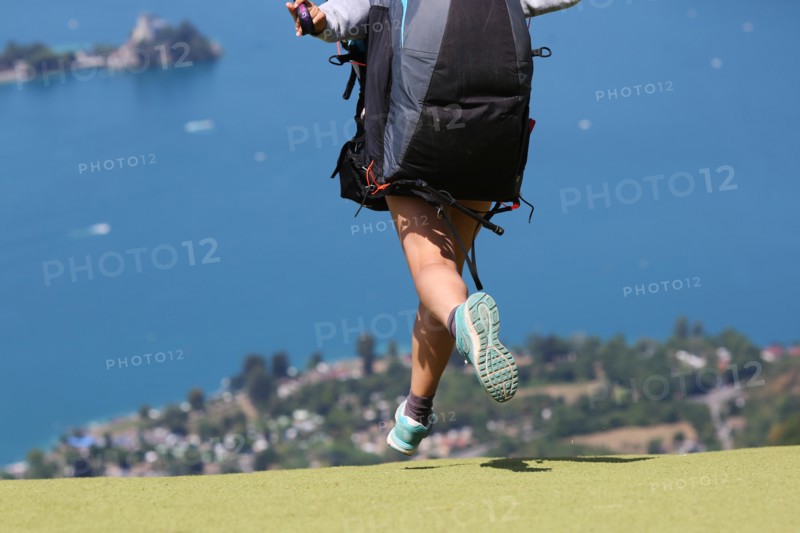 A female paragliding pilot taking off at Col de la Forclaz, near Annecy, France, with Lake Annecy in the background.