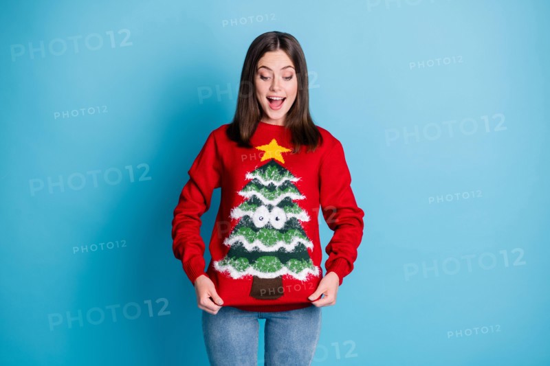 Photo portrait of surprised girl pulling ugly christmas sweater with hands smiling looking down isolated on pastel light blue colored background