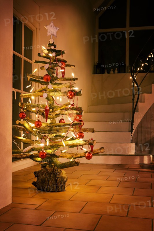 Rustic Christmas tree made of raw wood branches decorated with glowing fairy lights and red balls in a stairwell at night, selected focus, narrow dept