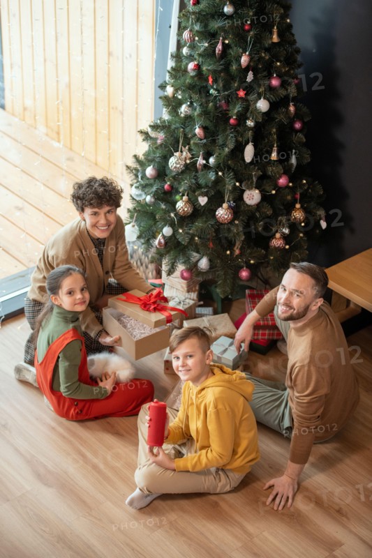 Portrait of positive beautiful Caucasian family sitting with presents on floor against Christmas tree at home