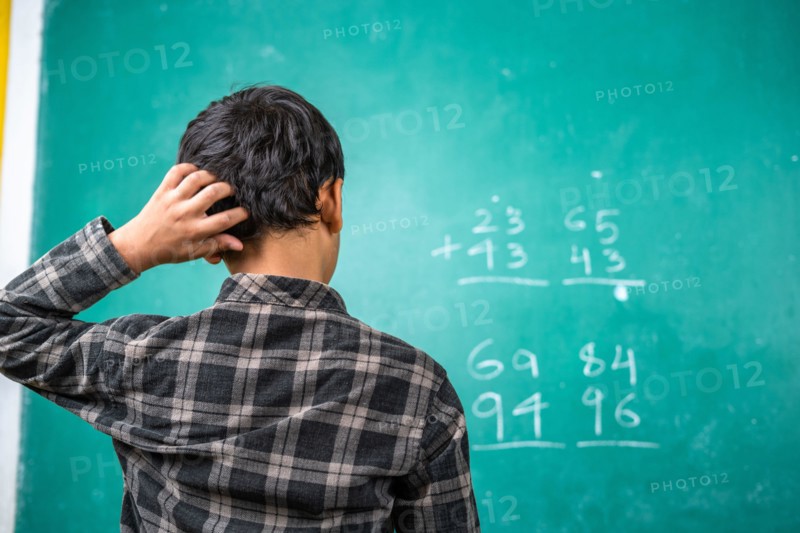 Confused kid scratching head to solve mathematics problem on board at classroom - concept of intelligence, trouble learning and education