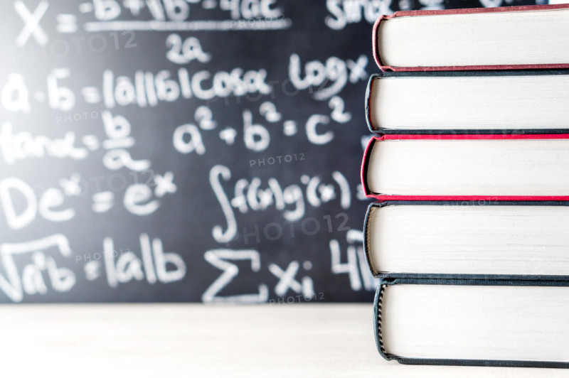 Stack and pile of books in front of a blackboard in school. Math equation handwritten on chalkboard.