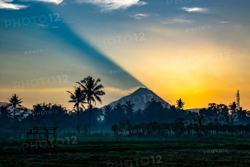 The casts a shadow as it rises over Mt Merapi, an active volcano in Central Java, Indonesia. Smoke spilling from the caldera.