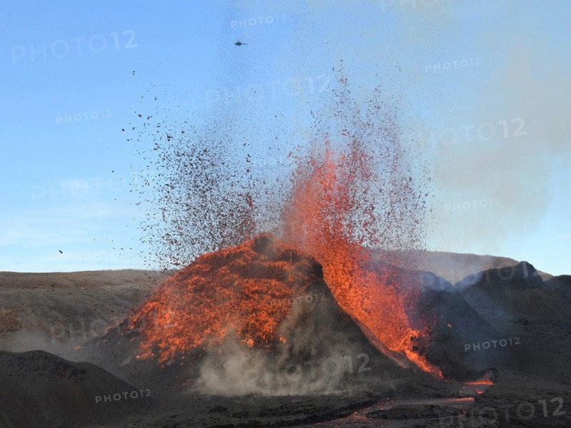 Volcanic vent at Fagradalsfjall, Iceland, erupting incandescent orange and red lava. Black lava in the foreground and blue sky in the background.
