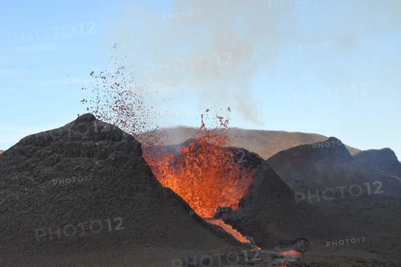 Volcanic vent at Fagradalsfjall, Iceland, erupting incandescent orange and red lava. Black cool lava in the foreground and blue sky in the background.
