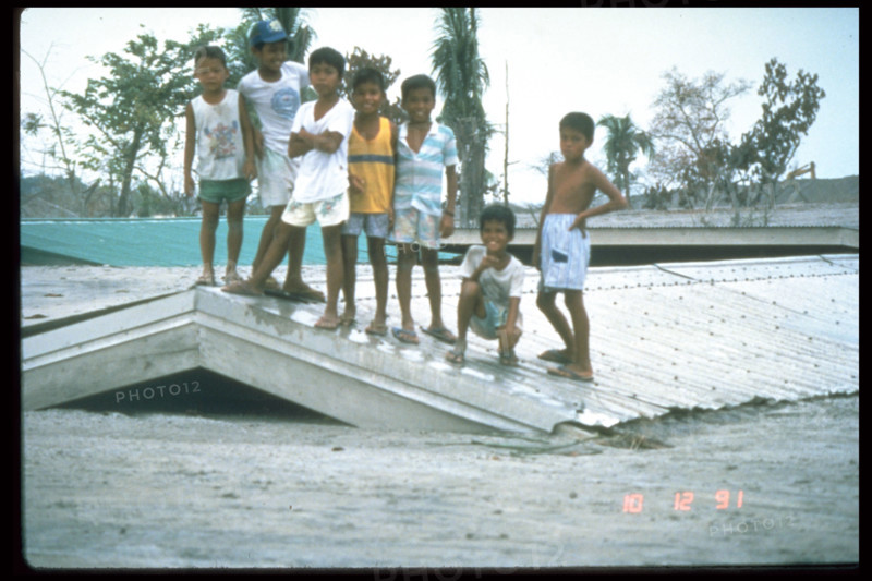 This photograph captures children on a roof in Bamban, Philippines, on December 10, 1991, during the aftermath of the catastrophic eruption of Mount Pinatubo. The eruption caused widespread destruction, with ash clouds affecting nearby areas.