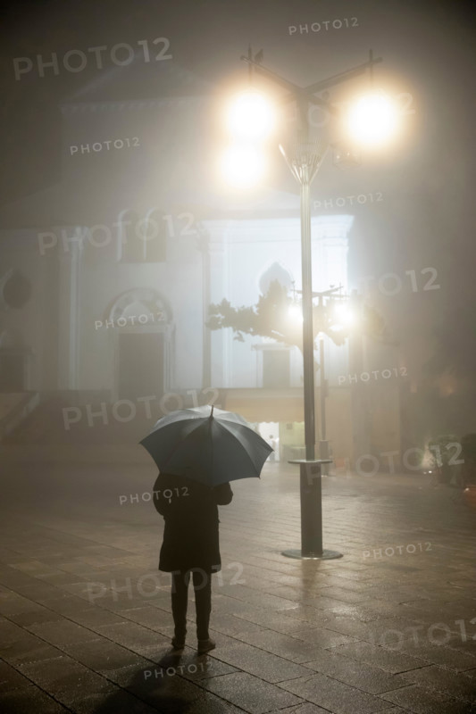 Person with umbrella standing under street lamp in front of the duomo in rain and fog at night, Ravello, The Amalfi Coast, Campania, Italy, Europe