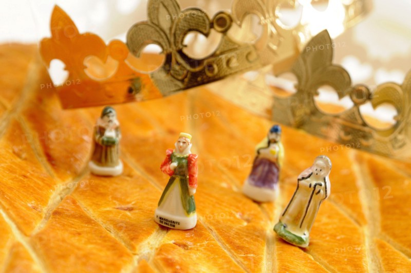 A golden, flaky galette des rois is on a yellow plate alongside traditional porcelain figurines and a paper crown. French Epiphanie
