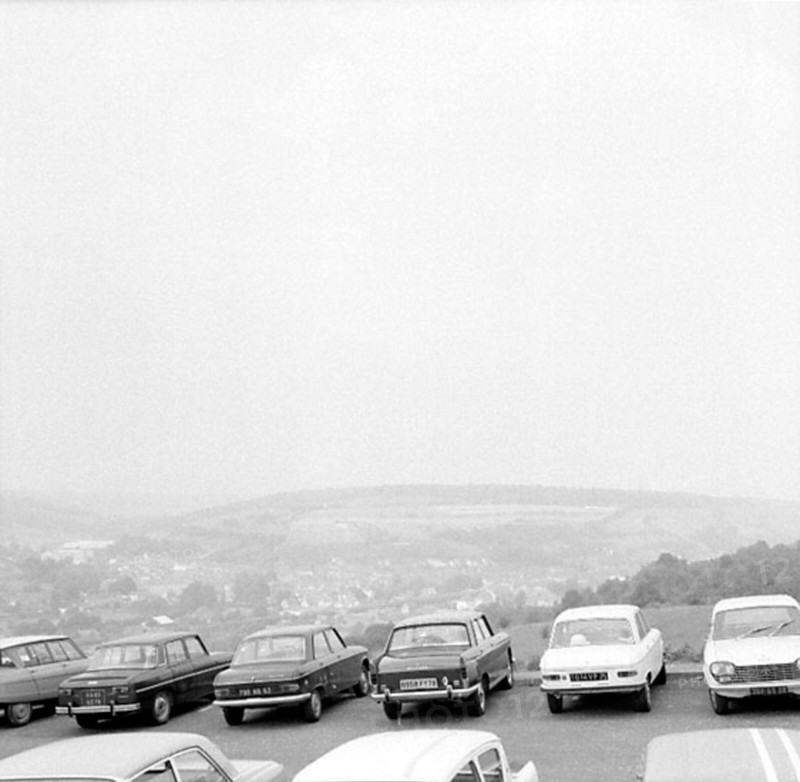 A number of cars are parked in a parking lot on a hill. Down in the valley is a community. Gandreas Uddling commented on the picture via the Digital Museum February 15, 2013: "View from France!" Car parking France 1960s.