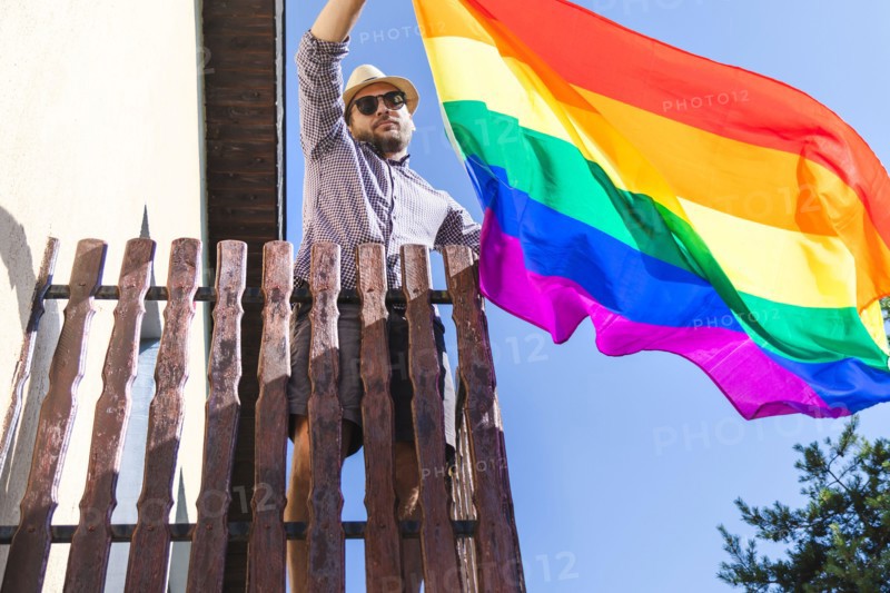 Man in hat holding lgbt rainbow flag in his hands while standing on the balcony.