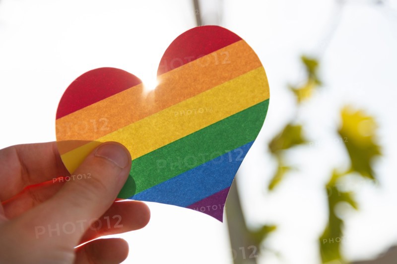 Hand holding a paper heart in LGBT rainbow colors with the sun rays shining around it, concept of LGBT, close-up