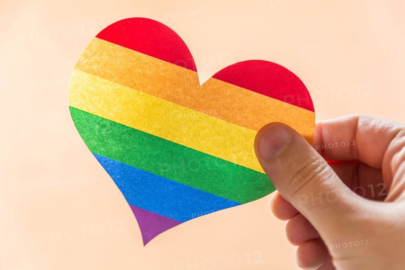 Hand holding a paper heart in rainbow colors or LGBT flags against a pink background, close-up