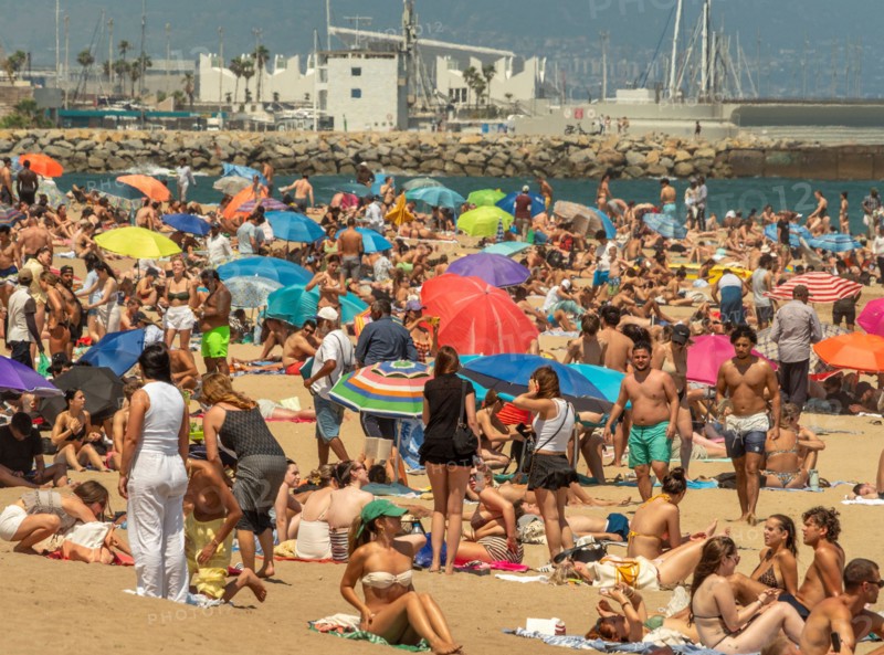 A bustling, crowded beach in Barcelona filled with people and colourful umbrellas, with the boats and breakwater of the port visible in the background.