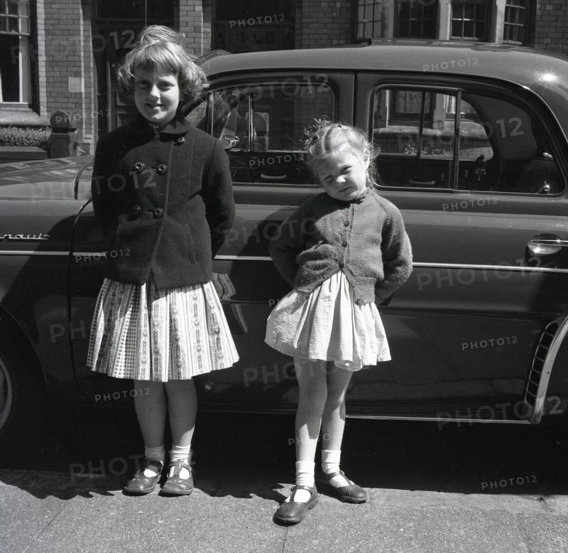 1958, historical, two young girls, sisters, stand outside in a British street by their father's car, a French Renault Dauphine, a rear-engined 4-door economy car, a successor to the Renault 4CV.  Between 1957 and 1961, Dauphines were assembled at Renault's Acton plant in West London and one was even driven HM the Queen. Roomy, with distinctive styling, the Dauphine car was a global success for Renault.