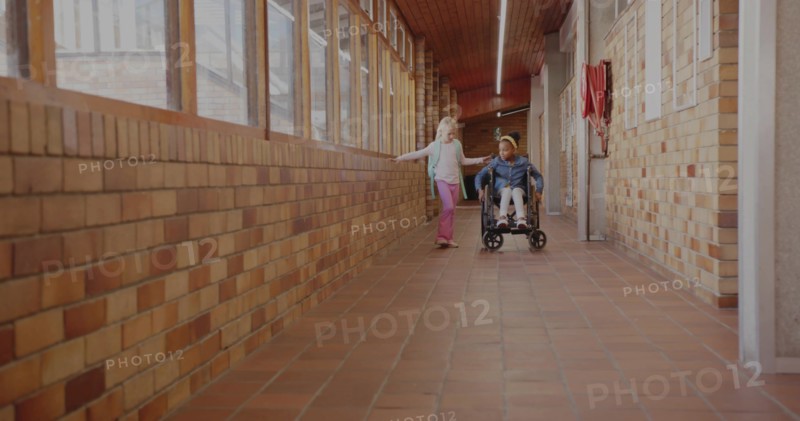 Two classmates rolling wheelchair and walking with backpack in school hall, with fire hose cabinet
