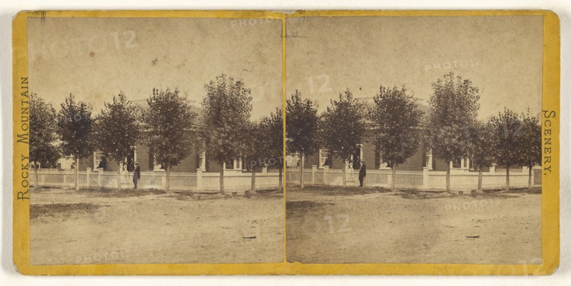 Rocky Mountain Scenery. Man in hat standing in front of a large tree-lined house, with white picket fence; American; about 1870