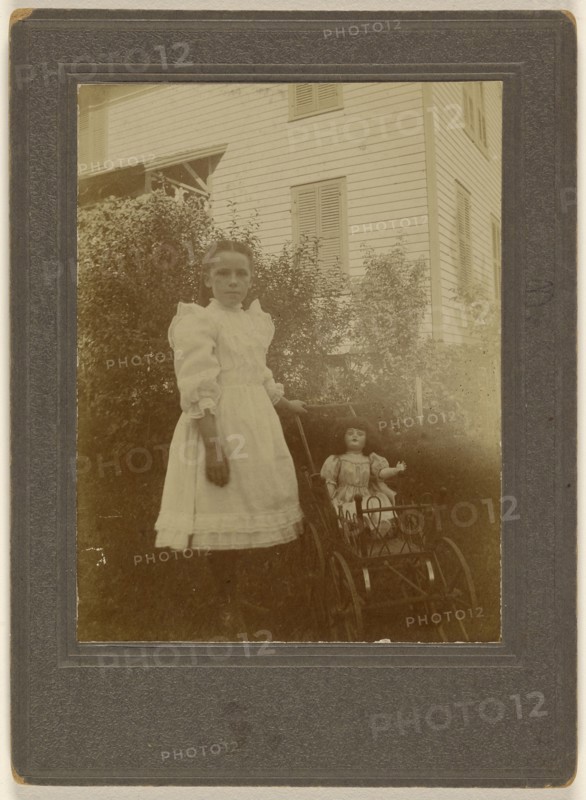little girl in white dress standing next to a baby carriage with a doll inside; American; about 1900; Gelatin silver print