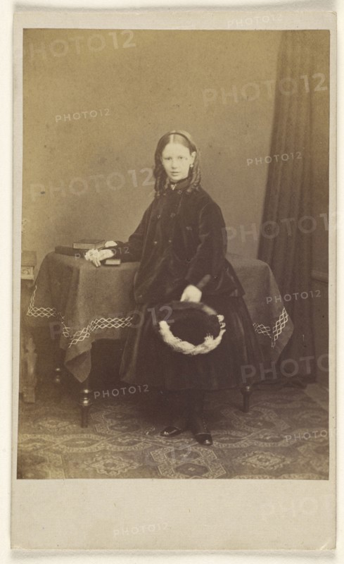 young girl in curls, standing, holding a hat, resting her other arm on a table with books on top; Joseph Brown, British, active