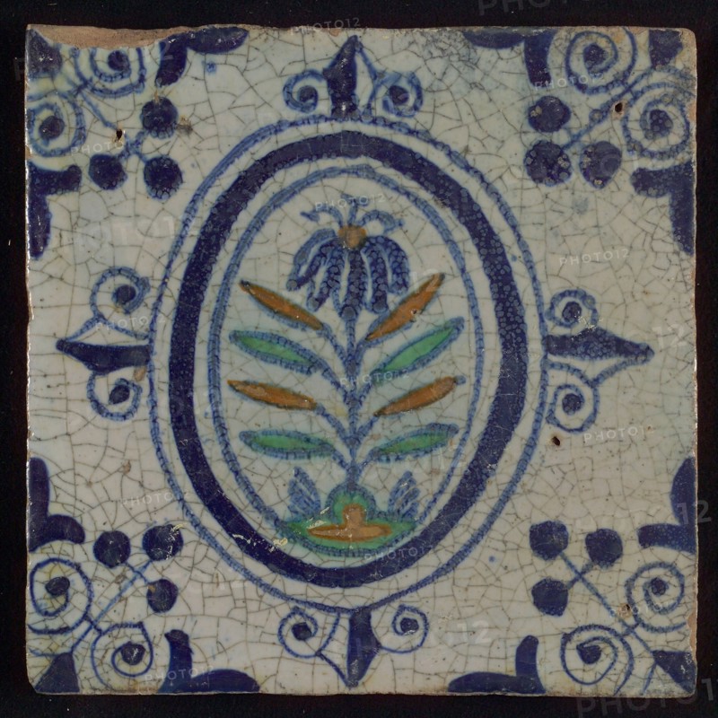 Tile, flower on spot in blue, green, and orange on white, inside an oval with lilies, corner motif, wall tile tile sculpture