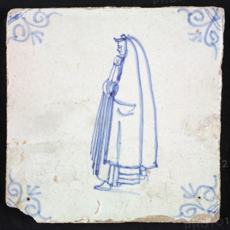 Figure tile, blue with civilian woman with hood, cloak that reaches from the head almost to the ground, corner motif oxen head