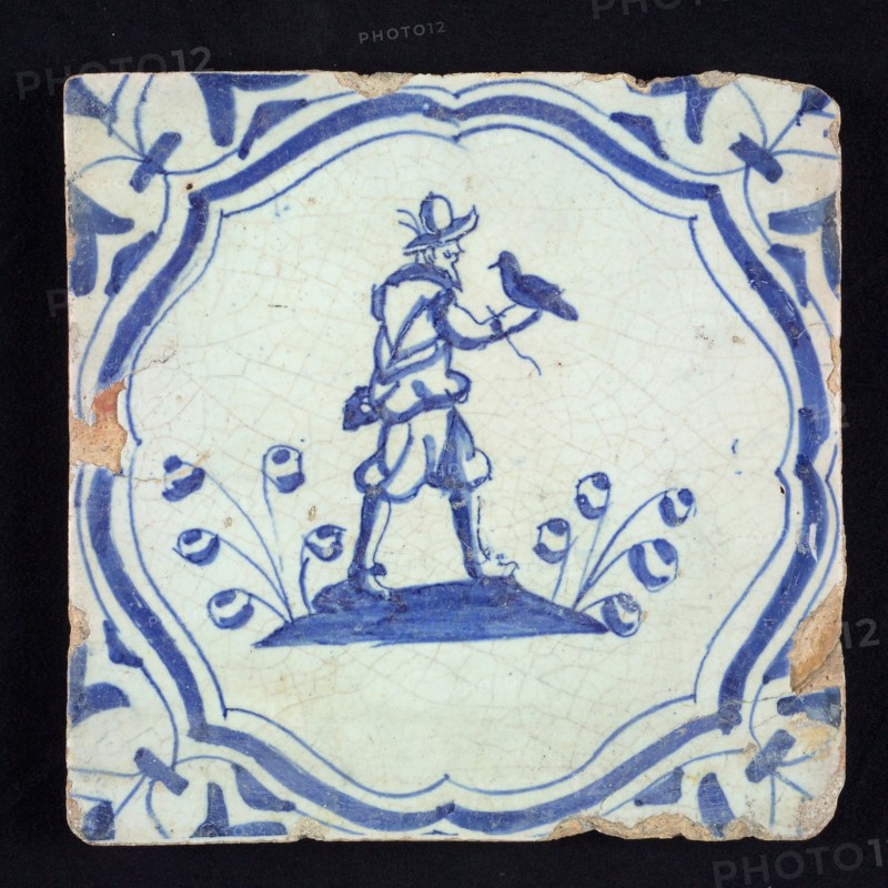 Figure tile, blue with falconer with falcon on the right hand, on the ground, in curly braces with corner motif, wall tile
