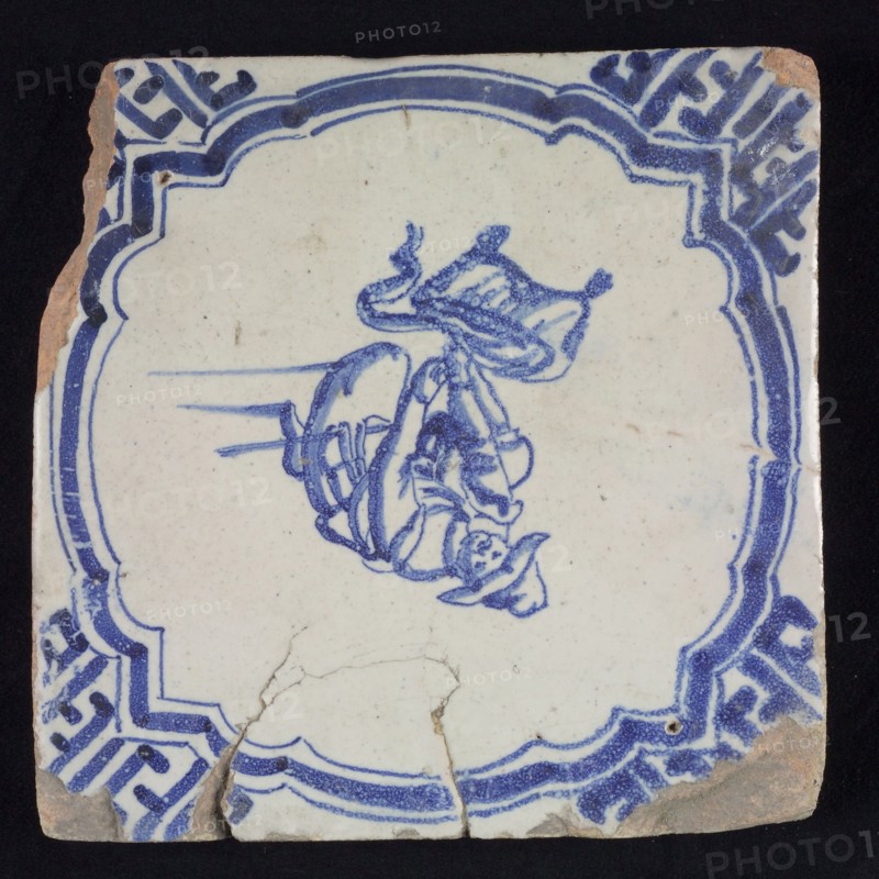 Figure tile, blue with man on knees, hunter taking something out of his bag, scalloped frame with braces, corner motif meander