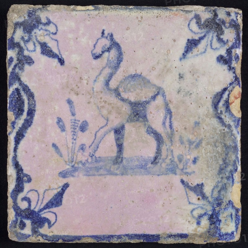 Animal tile, standing dromedary to the left on plot between balusters, in blue on white, corner pattern french lily, baking