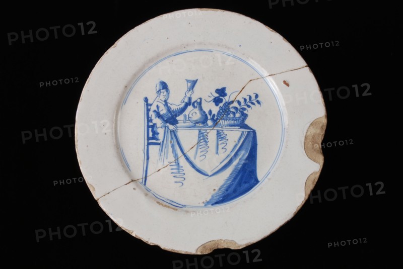Faience plate on stand, blue on white, man with raised glass on covered table, plate crockery holder soil find ceramic