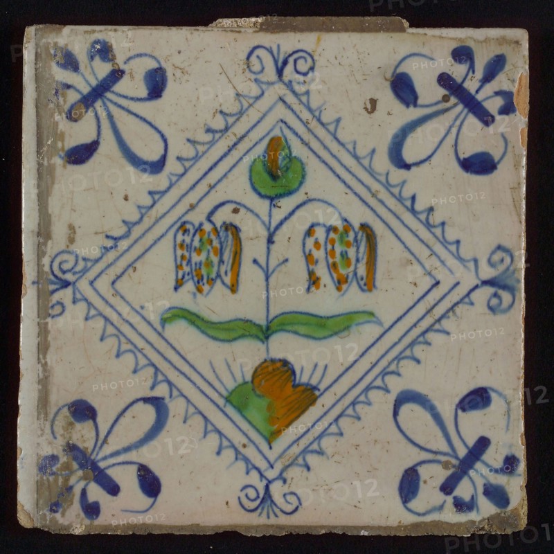Tile, double flower on ground in orange, green and blue on white, inside serrated square with plume, corner pattern french lily