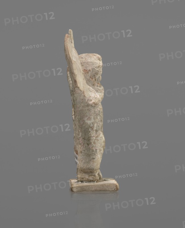 Terracotta figurine of Psyche. Young Psyche with butterfly wings, wrapped in a chlamys. Right hand rests on the chest, left hand rests along the body. Long hair falling on the shoulders arranged in curls. Head turned to the left. Figurine placed on a rectangular base. 332–30 BC, Hellenistic period; Tanagra Greece. Figurine; height 10.8 cm, width 7.5 cm, depth 3 cm., A small weathered figurine standing on a rectangular base, shown in profile with one arm raised; the surface appears rough and pitted with patches of worn material, and the form has subtle contours suggesting a head, torso, and limbs. The visible colors are pale beige, off-white, muted gray, and hints of light brown with some faint pinkish tones. The background is a flat medium gray and the base sits on a reflective surface that mirrors the bottom edge of the base.