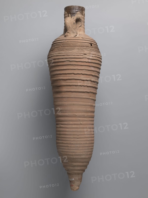 Amphora, unknown Egyptian workshop; wine transport vessel with a strongly elongated belly, narrowed in the middle. Roller-shaped vices, approximately oval in cross-section, are attached to the central section of the neck with a straight rim and connect to the upper part of the convex, steep shoulders. Conical base. Surface brown and even, covered with grooves of varying density and profile. Interior impregnated with a black resinous substance, with streaks visible in the upper neck. Clay brown, alluvial Naqlun AC1A ceramic body type. Inscription in red paint on the upper part of the handles: ICX. Hollowed-out hole ~0.5 cm diameter on the lower part of the handles below the inscription. 2nd half of the 4th–5th century; findspot Edfu. Vessel type: amphora. Dimensions: height 63.6 cm; rim diameter 6.8 cm; body diameter 19.3 cm., An elongated clay vessel with a narrow neck and a pointed base hangs against a pale gray background; the vessel has a ribbed texture formed by a series of horizon...