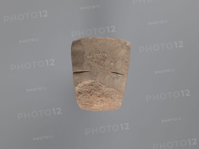 Pyramidal clay seal with a roughly square stamp surface and rounded corners. Shallow incisions at mid-height. Base engraved with a letter-like motif—either a geometric B or an M resting on the base—beside a small round recess. Upper part is impressed; chippings on the base. Visible kaolin clay, pink-beige. 6th–7th century, unknown South Egyptian workshop; Edfu. Seal/stamp. Height 6.1 cm, width 5.5 cm. Geometric decorations, monograms, engraved decorations, stamp impressions., A roughly rectangular stone object with a worn, textured surface suspended against a plain gray background; the stone shows earthy tones of brown, tan, and reddish-brown with rough, pitted areas and smoother, flatter areas; there are two shallow horizontal grooves near the upper middle and a more eroded, uneven lower portion with granular texture; overall the lighting highlights the stone’s surface irregularities and subtle color variations.