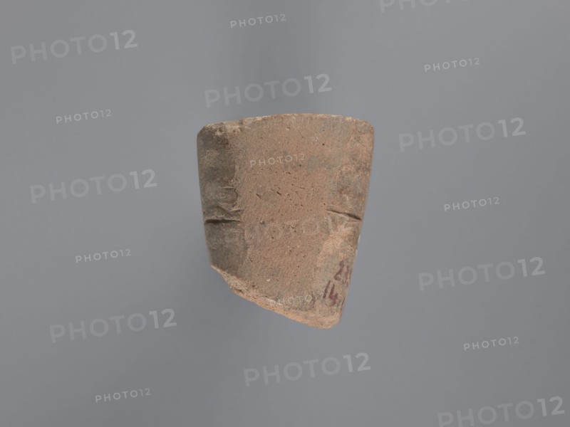 Seal, unknown South Egyptian workshop; pyramidal clay seal with a roughly square stamp surface, rounded corners; shallow incisions at mid-height; on the base an engraved letter possibly a geometrical B or an M resting on the base with a small round recess beside it; upper part is impressed, chippings on the base; visible kaolin clay, pink-beige; 6th–7th century; Edfu; seals, geometric decorations, monograms, engraved decorations, stamp impressions, stamps; seal/stamp; height 6.1 cm, width 5.5 cm, A roughly trapezoidal fragment of pottery floats against a neutral background. The fragment has a coarse, textured surface in shades of brown and tan with subtle darker brown areas and small speckles. The edges are worn and irregular, with a chipped corner showing a slightly lighter interior. There are a few shallow incised lines on the fragment’s face and faint darker smudges near one edge. The background appears as a smooth expanse of muted gray.