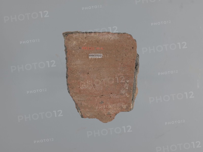 Fragment of a utility vessel. Made of type C Nile clay, highly porous with organic admixtures chaff and gold mica flakes. Thick fracture, dark gray Unworked surface exterior and interior brown, Engraved and plastic decoration, hand-applied. A horizontal half-cylinder, plastically modeled with recesses made with fingers or a round tool, separates the upper from the lower part. Top: a row of parallel recesses additionally decorated with a knurl. Lower: two partially preserved finger-shaped arcades made of glued-on clay rolls; a knob made from a clay disc is glued under one arcade. Wavy engraved lines run along the arcades. Above the arcades is a horizontal row of diagonal recesses—remains of the rope imprint used to tie the vessel during drying. 6th–7th century. Tell Atrib. Vessel; height 14 cm, width 11 cm, thickness 2 cm., A single irregular fragment of a hard material with a rough texture is centered against a plain background. The fragment shows a flat face with visible horizontal st...