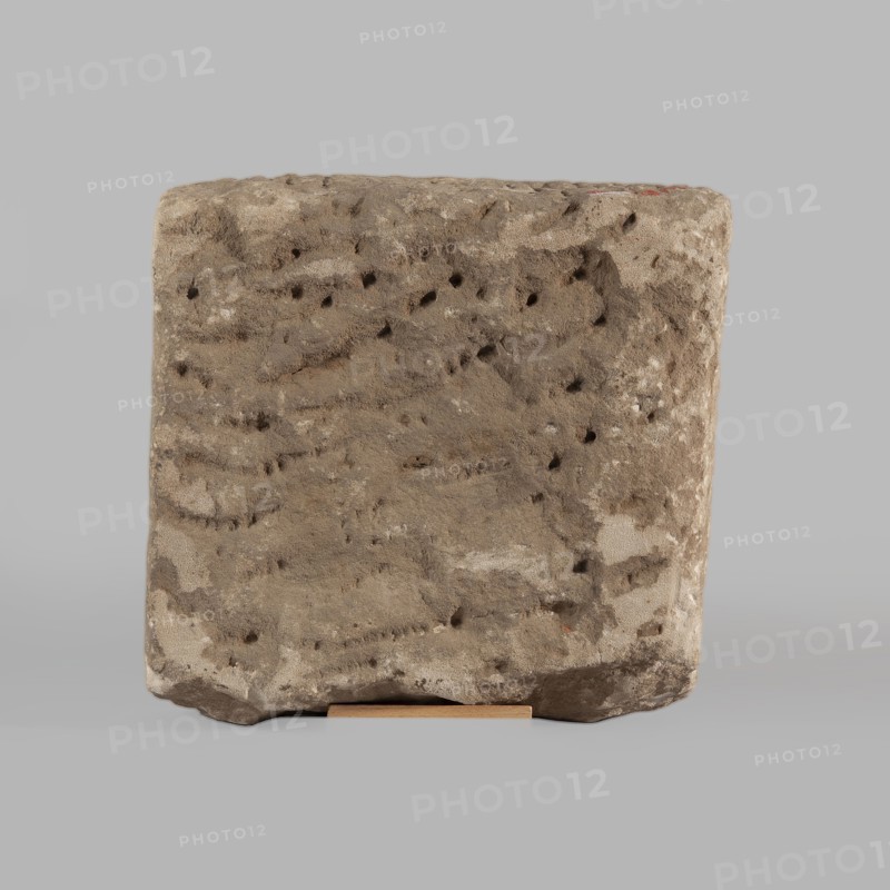 Fragment of a stele with an inscription in Arabic, unknown creator scribe-copyist, excavations, Islamic art, Arabic culture, inscriptions, Arabic inscriptions, Kufi script, Quran, Arabic language, Nubia, Nubian crafts. The surface of the block is smoothed; the inscription in Kufi script contains a fragment of the Quran 67:1. It is probably a funerary stele. 2nd half of the 11th–1st half of the 14th century. Origin: Faras, Sudan. Stele; height 32.0 cm, width 34.0 cm, depth 12.0 cm., A roughly rectangular block with a rough, pitted surface sits centered against a uniform light gray background, the block showing a textured, weathered appearance in shades of brown, beige, tan, and hints of darker brown in small pits and indentations; the top and sides display irregular grooves and a few lighter patches, while a thin flat support in a light wood color is visible under the block at the front.