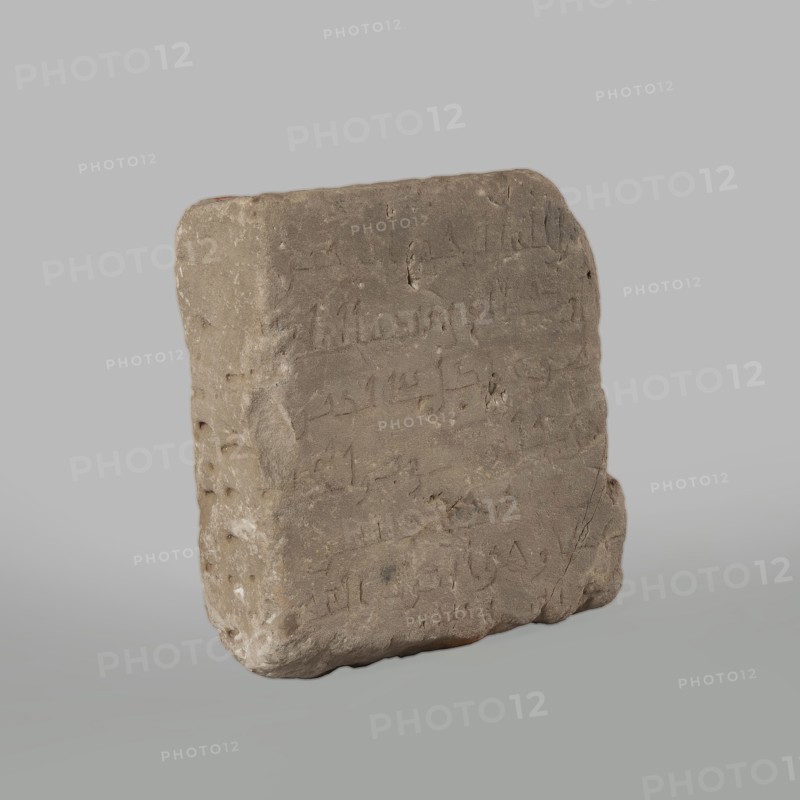 Fragment of a stele with an inscription in Arabic, excavations, Islamic art, Arabic culture, Arabic inscriptions in Kufi script containing a fragment of the Quran 67:1. Probably a funerary stele. Surface smoothed. 2nd half of the 11th–1st half of the 14th century. Origin: Faras, Nubia Sudan. Stele; height 32.0 cm, width 34.0 cm, depth 12.0 cm., A roughly rectangular stone slab with rounded edges and an uneven surface, shown standing against a plain light gray background. The slab is a dusty brown-gray color with areas of slightly lighter and darker brown, and small patches of pale off-white on the sides. The front face shows shallow incised marks and irregular indentations across its surface. The left side has a vertical row of rounded hollows and some textured roughness, while the right edge is a bit more worn and chipped. The top edge is uneven with a subtle curve, and the bottom edge is slightly thicker and irregular, allowing the slab to rest upright. The overall appearance is matt...