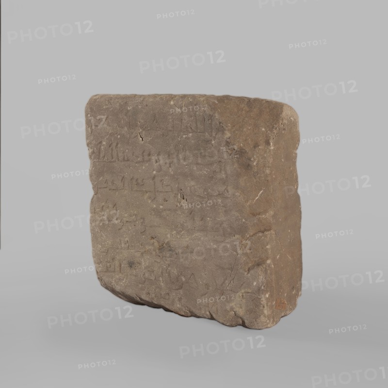 Fragment of a stele with an inscription in Arabic, unknown creator/scribe, excavations, Islamic art, Arabic culture, Kufi script, contains a fragment of the Quran 67:1, probably a funerary stele, 2nd half of the 11th–1st half of the 14th century, Faras, Sudan; stele; smoothed surface; height 32.0 cm, width 34.0 cm, depth 12.0 cm, A roughly rectangular stone slab stands upright against a pale gray background, showing an uneven surface with weathered textures, small chips, and visible cracks. The slab displays shallow incised marks across its face and subtle irregularities along its edges, with one rounded corner and a slightly tapered side. The visible colors include shades of brown ranging from light tan to darker brown, small areas of gray, and the surrounding backdrop in pale gray.