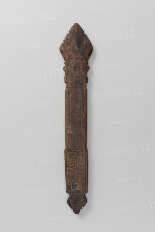 Hand cross, Diamond-shaped arms, wide grip almost same width as the arms, merging into a poorly defined plaque in shape of an elongated rectangle topped with a trefoil inset. Bilateral decoration, almost identical on sides A and B, limited to simple incisions at the edges of the cross arms and horizontal zigzag stripes and a double X on the plaque. 16th–20th century, Ethiopia. Metal cross, height 28 cm, width 5 cm., A long, narrow carved wooden object with a dark brown, weathered surface against a light gray background; the top end is shaped with a pointed, scalloped outline and the bottom end tapers to a decorative, rounded tip with carved geometric motifs near the lower section; the central shaft is relatively smooth and rectangular, showing natural wood grain and worn areas, and the lower portion features incised patterns with zigzag and cross-hatched textures.