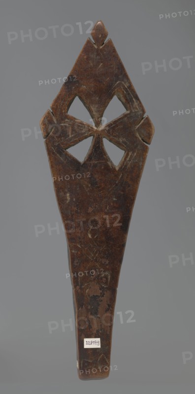 Hand cross fragment, Simple engraved decoration on the surface: four triangular cutouts arranged in the form of a cross; below the holes are two rather carelessly engraved crosses. The reverse is also decorated with a cross motif; decoration engraved with a thin, shallow line. Dark wood. 20th century, Ethiopia. Cross; height 26 cm, width 8 cm., A vertically oriented carved wooden object with a dark brown surface showing lighter worn areas and scratches, featuring a pointed top with a small raised diamond-shaped element and carved cutouts forming a symmetrical four-petaled motif near the upper portion, flanked by small protruding shapes on each side; the lower portion tapers into a handle-like shape with visible patches of lighter and darker brown wear and some faint carved line patterns, all set against a neutral gray background with a few small areas of lighter discoloration on the wood.