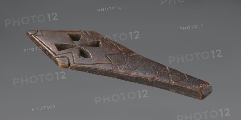 Hand cross - fragment. Simple engraved decoration on the surface; four triangular cutouts arranged in the form of a cross. Below the holes, two rather carelessly engraved crosses are visible. The reverse is also decorated with a cross motif. Decoration engraved with a thin, shallow line. Dark wood. 20th century, Ethiopia. Cross; height 26 cm, width 8 cm., An elongated carved wooden object with a tapered end and a wider opposite end, showing a carved geometric cutout near the wider end and shallow incised linear decorations along its surface; the wood surface displays worn and weathered areas with a patina ranging from dark brown to lighter brown and small patches of black, set against a plain grey background.