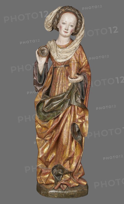 Figure of St. Mary Magdalene, ca. 1480, Southern Germany. Hollow wall sculpture, height 115.0 cm, width 35.8 cm, depth 16.2 cm. St. Mary Magdalene, depicted frontally in slight contrapposto with her head tilted left, holds a perfume box in her left hand and its lid in her right. The figure’s movement and hand gestures are elegant. The face has small, delicate features and a high forehead, framed by a thick braid woven with a ribbon; a white shawl draped over her head falls left, covers the breast, and is draped over the right shoulder. She wears a double gown: an over-dress of patterned golden fabric, short-sleeved and tied at the waist creating shallow radiating pleats, and an under-dress originally dark red, now darkened visible in the flared sleeves. A cloak is draped over the left shoulder, its pleated tail draped over the front and held by the left hand, revealing a green lining where folded back; on the right it forms deep, relatively stiff folds with semicircular and V-shaped ou...