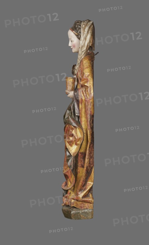 Figure of St. Mary Magdalene, ca. 1480, Southern Germany. Hollow wall sculpture, height 115.0 cm, width 35.8 cm, depth 16.2 cm. St. Mary Magdalene is depicted frontally in a slight contrapposto with her head tilted left; in her left hand she holds a perfume box and in her right its lid. The figure’s movements and hand gestures are elegant. The face has small, delicate features and a high forehead, framed by a thick braid woven with a ribbon; a white shawl draped over her head falls to the left, covers the breast and is draped over the right shoulder. She wears a double gown: an over-dress of patterned golden fabric, short-sleeved and tied at the waist creating shallow radiating pleats, and an under-dress originally dark red, now darkened visible in the flared sleeves. A cloak is draped over the left shoulder, its pleated tail draped over the front and held by the left hand, revealing a green lining where folded back; on the right the cloak forms deep, rather stiffly modeled folds with ...