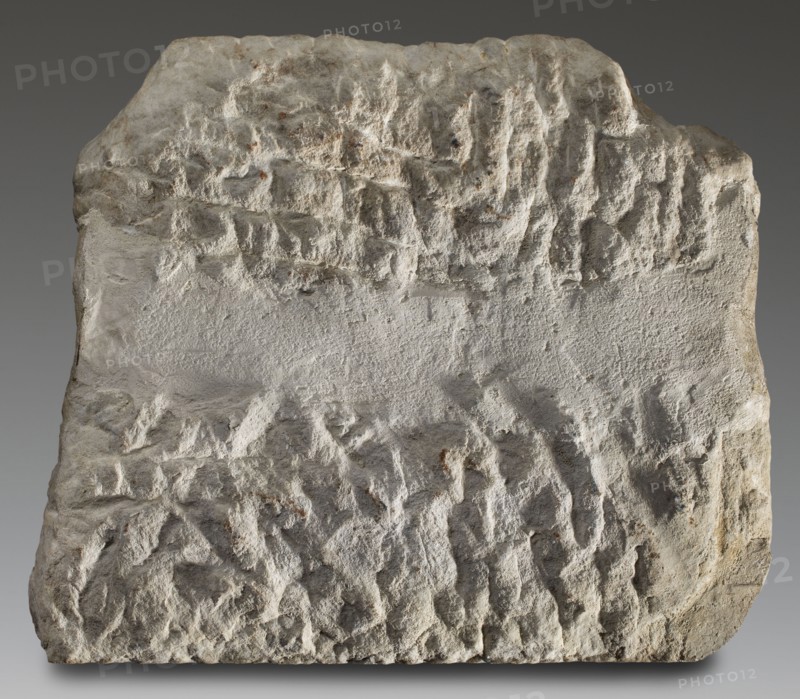 Honorific inscription, unknown workshop of Asia Minor author; Roman legions, military decorations; 120–130; Asia Minor Heraclea Salbake; inscription; height 57 cm, width 49 cm, depth 8 cm, weight 49.5 kg, A roughly rectangular block of stone with an uneven, weathered surface, showing a mix of smoother and heavily textured areas; the stone displays shades of gray and off-white with small patches of brownish rust coloring and subtle darker gray shadows in crevices; the top and lower portions have pronounced ridges and pitted indentations while a central horizontal band appears relatively smoother and slightly lighter; the edges are irregular and worn, and the overall appearance is matte and coarse.