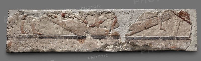 Fragments of wall decoration from the mastaba of the dignitary Tepemankh. Scenes of boat building and bringing funerary offerings. Old Kingdom, 5th Dynasty, reign of Niuserre c. 2445–2421 BC. Abusir. Bas-relief; height 26 cm, width 302 cm, depth 16 cm, weight 140 kg. Includes depictions of sacrificial artists, scribes, shipwright; funerary/sepulchral architecture; inscriptions naming Niuserre and Tepemankh., A rectangular stone relief with worn carved figures and a rough textured surface, showing a horizontal composition of reclining and standing human-like forms rendered in shallow incisions and low relief; the stone displays areas of smooth carving and pitted erosion, with visible remnants of painted areas in shades of red and brown, and traces of darker gray and black along a horizontal band; the overall stone color is off-white to beige with spots of darker discoloration and weathering, and the background behind the relief is a uniform medium gray.