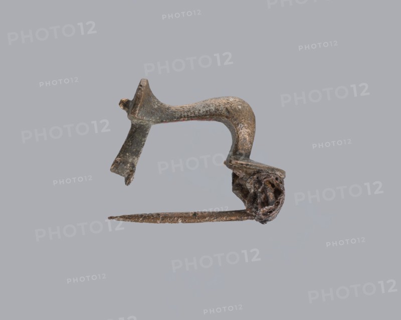 Knuckle-shaped fibula, unknown Roman workshop. Fibula with a flat underside, rounded at the top, rising from a horizontal semicircle upwards, bent at an acute angle and terminating in a semicircle with a button in the center. Below the arch, on one side, there is a long spiral, from which a pin emerges; on the other, a high stem at the end is bent into a sheath. Material: bronze. Condition: good. Date: 2nd half of the 2nd–1st half of the 3rd century. Culture: Roman Empire 27 BC–476. Type: fibula. Dimensions: height 2.6 cm, width 3.7 cm., A single small metal object with a curved upper section and an attached straight pin-like lower piece is displayed against a plain light gray background; the object appears corroded and rough with a patina showing dark brown, dull bronze, and hints of greenish tarnish, and there is a twisted, irregular cluster of metal at one end while the opposite end shows a flattened, broken-looking extension.