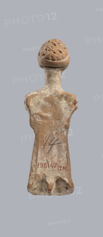 Terracotta figurine of a woman, terracotta, female molded figurine, Greece, orange clay, surface covered with whitewash. Woman dressed in a short, pleated chiton girdled at the waist. Long, thick neck and disproportionately small head with an oval face. Hair combed upwards and tied in a bun, held in place by a headband tenia tied in a bow at the top of the head. The back of the figurine is unfinished. The figurine originally had separately attached arms and legs below the knees, as evidenced by a recess and perforations for hooks. Condition: missing arms and legs, whitewash worn away. c. 400 BC, Greece, toy. Height 10.1 cm, width 3.5 cm., An upright worn figurine with a rounded textured head and a narrow neck leading to sloping shoulders and an elongated torso, displaying a weathered surface with patches of lighter and darker areas; the object shows rough, chipped edges and small pits, and the visible colors include shades of tan, brown, beige, and off-white against a plain gray backgr...