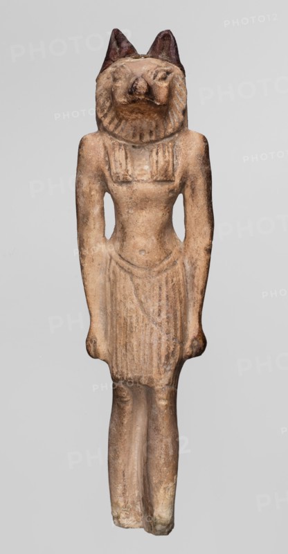 Amulet — Anubis. Anubis is depicted as a striding man with the head of a dog Canidae. Arms hang at his sides, hands clenched. Torso broad at the shoulders, narrowed at the waist. Pleated skirt with a loose belt, slightly lowered in the middle, reaches above the knees. Tripartite wig covers the head. A sort of mane is visible around the narrow, carefully modeled muzzle. Small, clumsily made ears are second-hand additions. Rear pillar reaches 2/3 of the figurine's height, with a drilled hole in its upper part. ca. 1069–525 BC; Third Intermediate Period–26th Dynasty. Origin: Egypt. Amulet. Height: 7.4 cm., A standing carved figure with an animal-like head and upright ears, depicting a human-shaped body wearing a short pleated skirt and a fitted upper garment, all in a weathered beige to light brown stone with darker brown accents on the ears and some shadowed areas; the surface shows rough texture and areas of lighter cream wear, with subtle variations of tan and ochre across the torso, a...